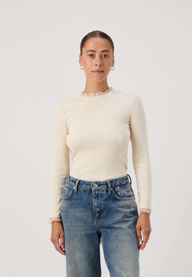 Long-sleeve cream ribbed top with a ruffled neckline, paired with high-waisted faded blue jeans. Simple design, soft texture.