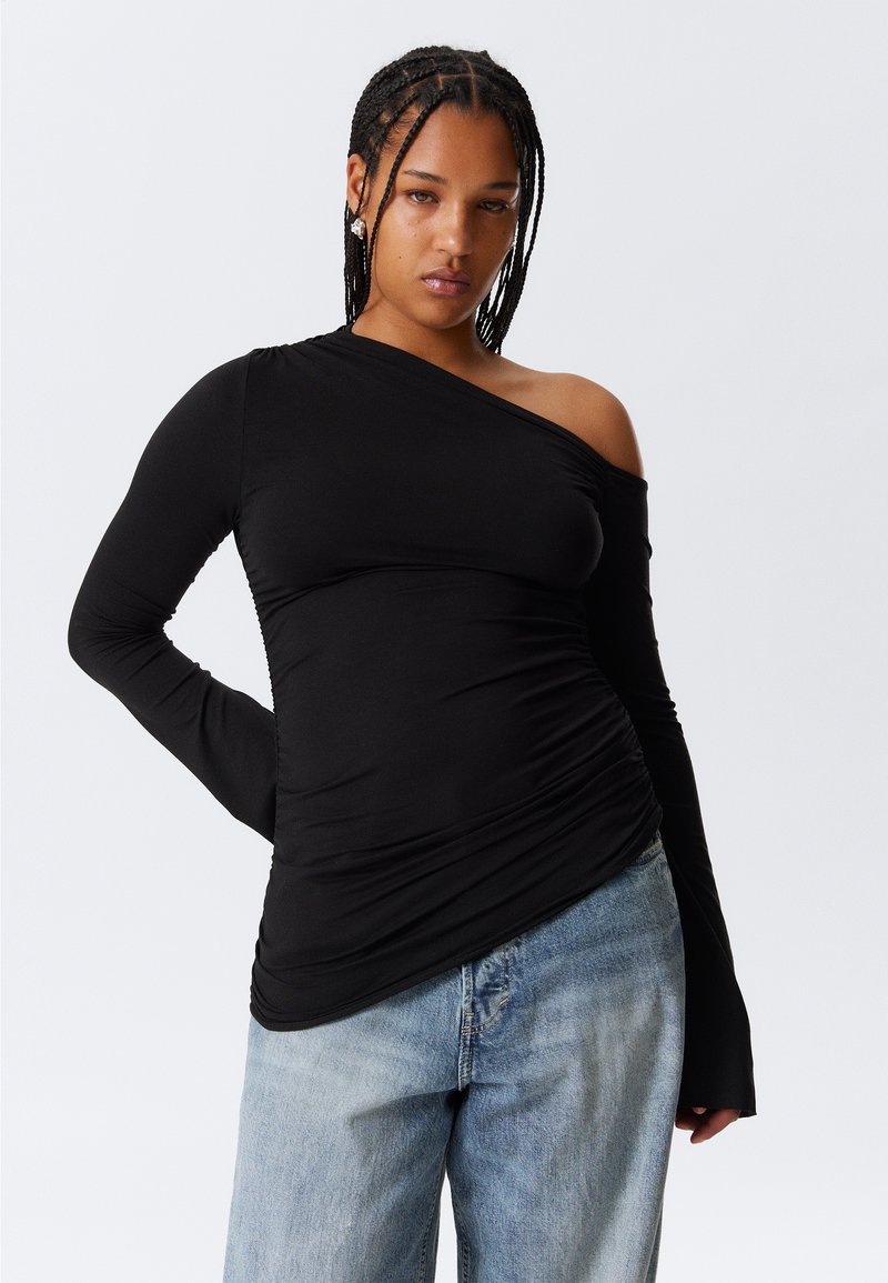 Black long-sleeve top with asymmetric neckline and gathered texture; fitted design contrasts with relaxed blue denim jeans.