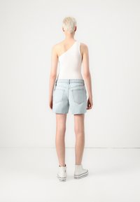 Shorts de ganga azul claro com a bainha desfiada, combinados com uma blusa branca de um ombro só. Pessoa a usar ténis brancos altos, vista por trás.