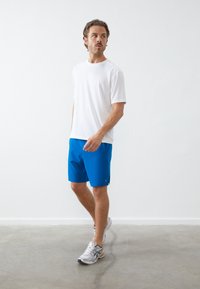 Λευκό κοντομάνικο T-shirt συνδυασμένο με μπλε αθλητικά σορτς και γκρι αθλητικά παπούτσια, σε απλό ανοιχτό φόντο.