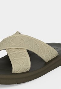 Sandale beige avec des brides croisées en tissu texturé et une semelle épaisse foncée, montrée en gros plan sur un fond blanc.