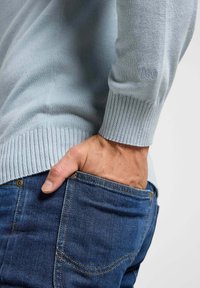 Lichtblauwe trui met geribbelde manchetten en onderrand, gemaakt van zachte stof. Zichtbaar stikseldetail op denim jeans, hand in de zak.