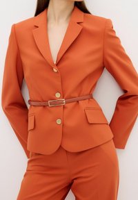 Femme portant un blazer ajusté orange brûlé avec des boutons dorés et une fine ceinture marron, associé à un pantalon assorti sur un fond uni.
