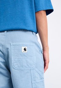 Svijetloplave cargo hlače imaju kvadratnu džepu s crno-bijelim Carhartt logotipom. Tkanina izgleda glatko s vidljivim detaljima šavova.
