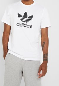 Bílý bavlněný tričko s velkým černým logem adidas a emblémem se třemi listy na přední straně; kulatý výstřih a krátké rukávy.