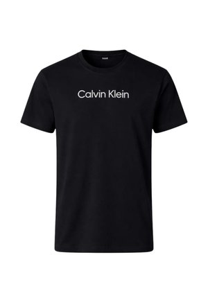 Giovane uomo che indossa una t-shirt nera Calvin Klein, jeans neri e scarpe nere, che tiene in mano una giacca scura piegata nella mano sinistra.