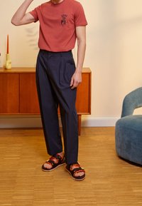 Camiseta roja de manga corta con logo, pantalones de tiro alto azul marino con pliegues y sandalias negras con acentos en rojo y naranja. Fondo de muebles de madera.