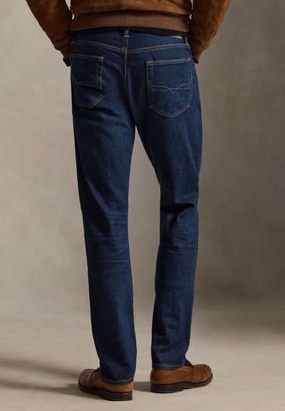 Dunkelblaue Jeans aus Denim mit geradem Schnitt, ausgestattet mit zwei Gesäßtaschen und dezenten Steppnähten. Kombiniert mit braunen Schuhen.