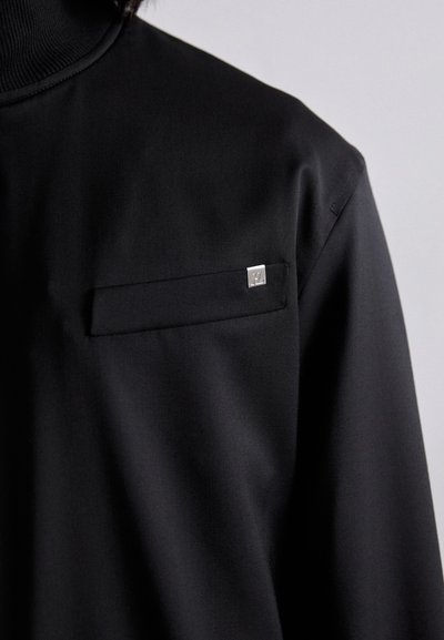 Black shirt with a high neck, featuring a small chest pocket and a silver hardware accent. Smooth texture, tailored fit.