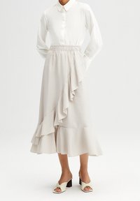 Falda beige claro con cintura elástica, detalle de volante diagonal, dobladillo acampanado y textura suave. Combinada con una camisa blanca.