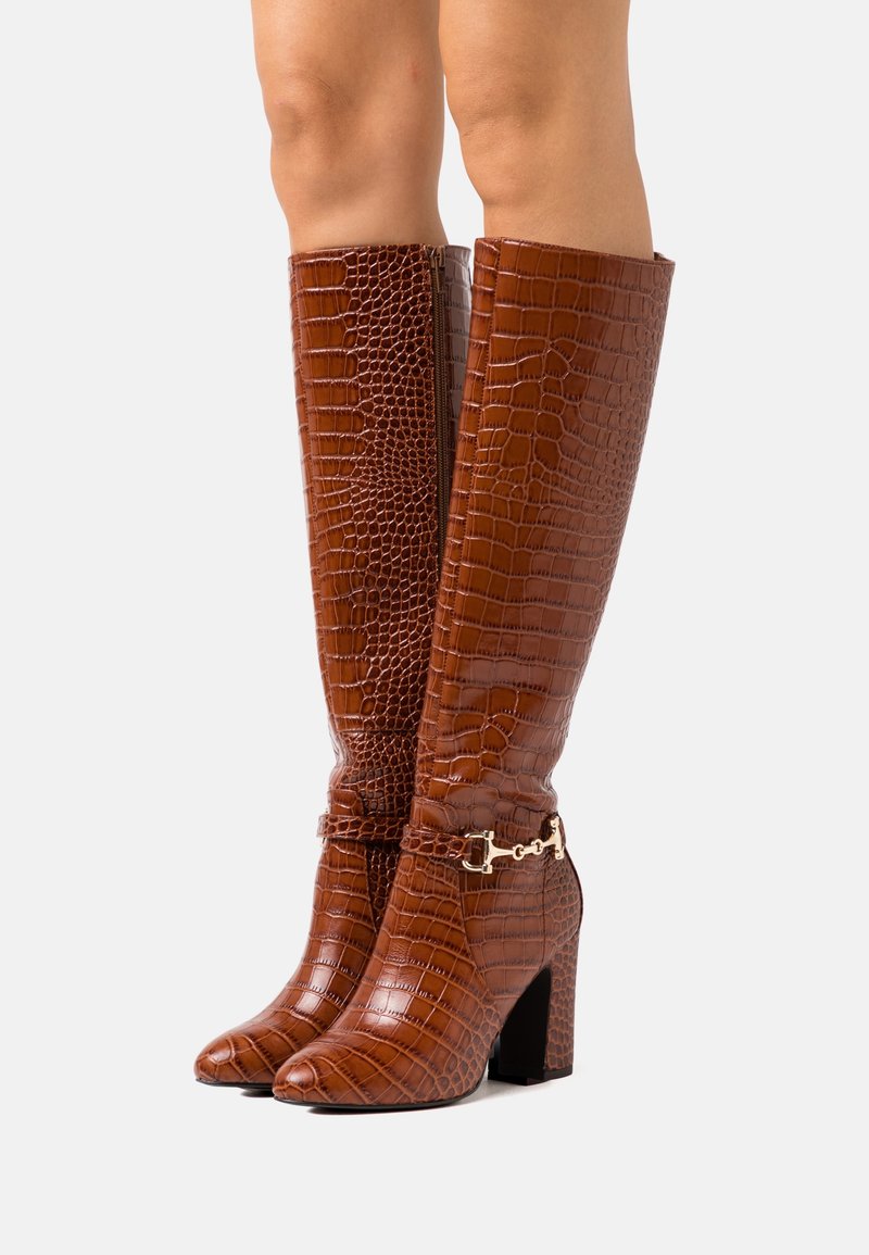 Bottes marron montant jusqu'aux genoux avec texture crocodile, talon bloc et détails en métal doré. Finition lisse avec une coupe ajustée.