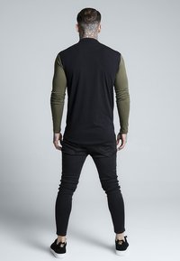 Black long-sleeve top with olive green sleeves, paired with black tapered jeans and black sneakers. Notable tattoo on the neck.
