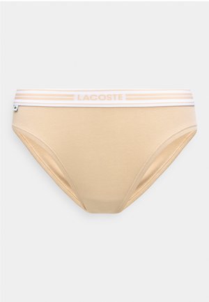Μπεζ γυναικείο μπικίνι σλιπ με λευκή ελαστική μέση που διαθέτει ανοιχτό μπεζ ρίγες και κείμενο "LACOSTE", καθώς και μικρή ετικέτα της μάρκας στο πλάι.