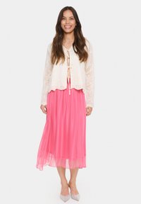 Cardigan corto in pizzo avorio con chiusura a cravatta sopra un abito maxi fluido rosa. Le scarpe sono a punta con dettagli scintillanti.