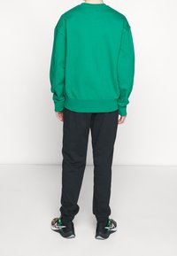 Person klädd i en grön sweatshirt, svarta joggerbyxor och svarta sneakers med färgglada mönster, står vänd med ryggen mot kameran på en enkel bakgrund.