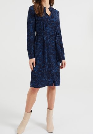 Frau trägt ein marineblaues, knielanges Hemdblusenkleid mit Blumenmuster, langen Ärmeln und gebundener Taille, kombiniert mit beigen Knöchelstiefeln.