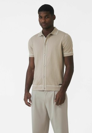 Beżowa koszulka polo z krótkim rękawem, z białym obszyciem i guzikami. Wykonana z gładkiej tkaniny, ma klasyczny kołnierzyk i prosty krój.