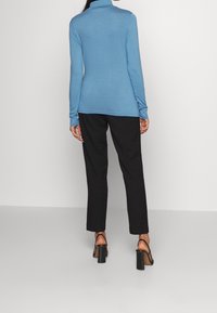 Ljusblå långärmad topp med hög krage, parat med svarta smala byxor och svarta högklackade sandaler, sett bakifrån.