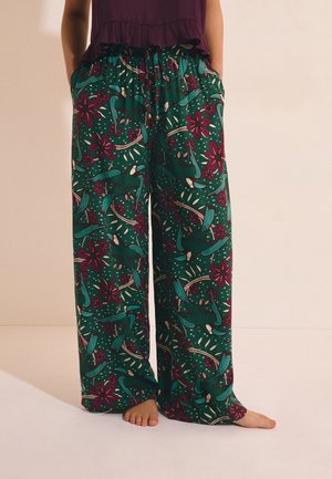 Zielone spodnie o szerokich nogawkach z turkusowymi ptakami i ciemnoróżowym kwiatowym wzorem, noszone na boso z rękami w kieszeniach i bordowym topem.