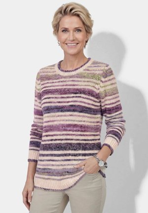 Vrouw met kort blond haar die een trui met paarse, roze en groene strepen en beige broek draagt, glimlachend met één hand in de zak.