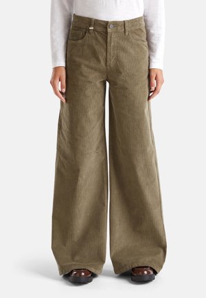 Henkilö pukeutuneena oliivinvihreisiin leveälahkeisiin corduroy-housuihin, joissa on etutaskut, ja ruskeisiin kenkiin, yhdistettynä valkoiseen pitkähihainen yläosaan.