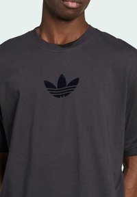 tmavě šedé bavlněné tričko s černým embosovaným logem Adidas na hrudi, kulatým výstřihem a krátkým rukávem.