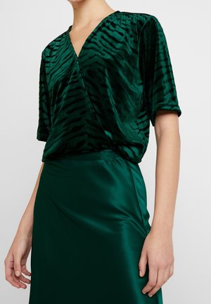 Femme portant un haut cache-cœur en velours vert à motif zèbre avec des manches longueur coude et une jupe assortie en satin vert.