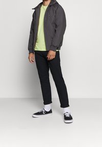 Hombre vestido con chaqueta con capucha gris y cremallera, camiseta verde claro, pantalones negros arremangados, calcetines blancos y zapatillas negras con cordones, de pie contra una pared blanca.