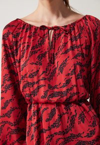 Red dress with black leaf pattern, elastic neckline, tie details, and bell sleeves. Soft fabric with a flowing silhouette.