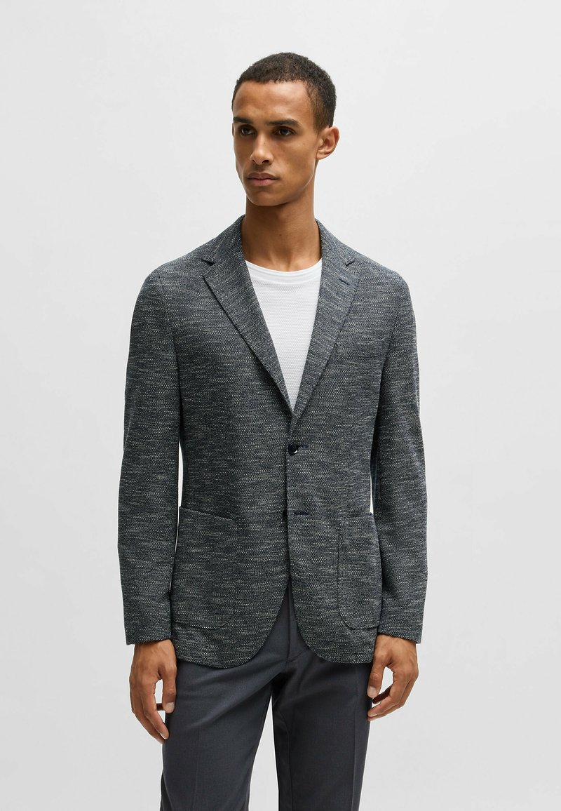 Blazer grigio texturizzato con un bottone singolo, due tasche frontali e rever a punta, abbinato a una T-shirt bianca e pantaloni scuri.