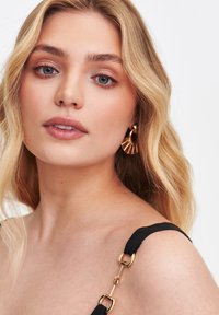 Młoda kobieta z długimi, falującymi blond włosami i delikatnym makijażem, ubrana w czarną bluzkę z złotymi detalami na ramiączkach oraz dużymi złotymi kolczykami.