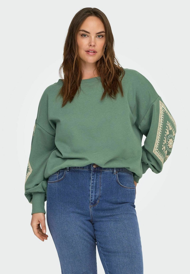 Felpa verde con maniche oversize a motivo, abbinata a jeans blu a vita alta. Il tessuto sembra morbido e dalla vestibilità comoda.