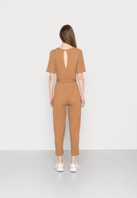 Brun ribbad jumpsuit med korta ärmar, figursydd midja och cut-out detalj på baksidan, kombinerad med vita sneakers. Slät textur, avslappnad design.