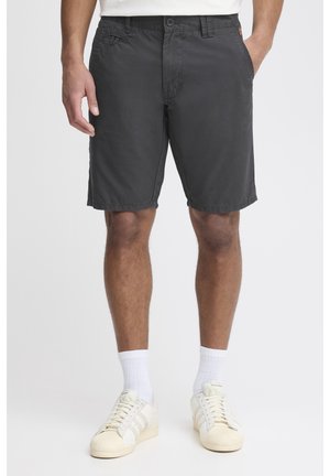 Homme portant un short gris foncé mi-cuisses, des chaussettes blanches montantes et des baskets blanc cassé, debout les mains dans les poches.