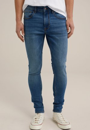 Slim-fit siniset farkut haalistuneella pesulla, ommeltu saumoilla, viisi taskua ja klassinen nappi- sekä vetoketjukiinnitys. Täydentävät valkoiset lenkkarit.