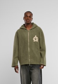 Hanorac cu fermoar de culoare verde măsliniu, cu glugă mare, buzunar frontal și logo brodat de culoare crem. Textură moale și croială lejeră.