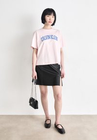 Rozā kokvilnas t-krekls ar zilu "CALVIN KLEIN" logo, apvienots ar melnu ādas mini svārku ar sānu šķēlumu un melnām plakanu apavu.