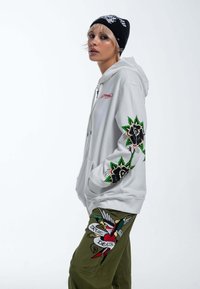 Sudadera blanca con un gran bordado de flores negras en las mangas, combinada con pantalones verde oliva que presentan coloridos diseños y patrones bordados.