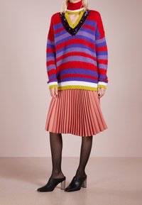 Kobieta ubrana w czerwono-fioletowo-żółty sweter w paski z cekinowym dekoltem w kształcie litery V oraz różową plisowaną spódnicę, czarne rajstopy i czarne mule na obcasie.