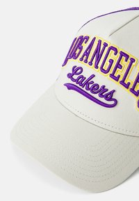 Beige baseballkeps med lila och gula broderade "LOS ANGELES" och "Lakers"-text. Har en böjd skärm och textilmaterial.