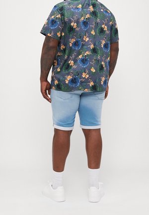 Homme portant une chemise à manches courtes à fleurs sombre, un short en jean bleu clair retroussé, des chaussettes blanches et des baskets blanches, debout de dos.