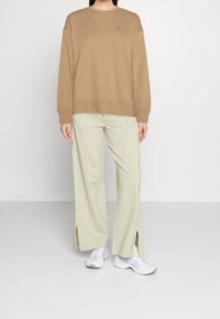 Brązowy, oversize'owy sweter z okrągłym dekoltem, w połączeniu z jasnobeżowymi spodniami typu wide-leg z bocznymi rozcięciami. Całość uzupełniają białe buty sportowe.