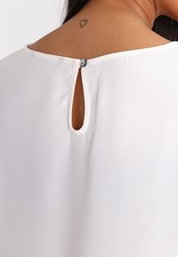 White blouse featuring a keyhole opening at the back, secured with a small button. Soft, smooth fabric with subtle sheen and minimal seams.