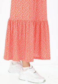 Korallfarbenes Kleid mit weißen Blumenmustern, которое с волнообразным подолом. Сочетается с белыми кроссовками, демонстрирующими массивный дизайн и сетчатые Akzente.