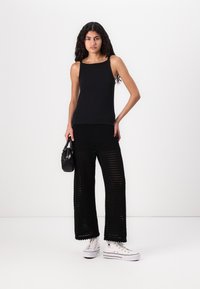 Top aderente nero abbinato a pantaloni larghi all'uncinetto neri. Sneakers bianche con dettagli neri. La modella tiene una piccola borsa nera.