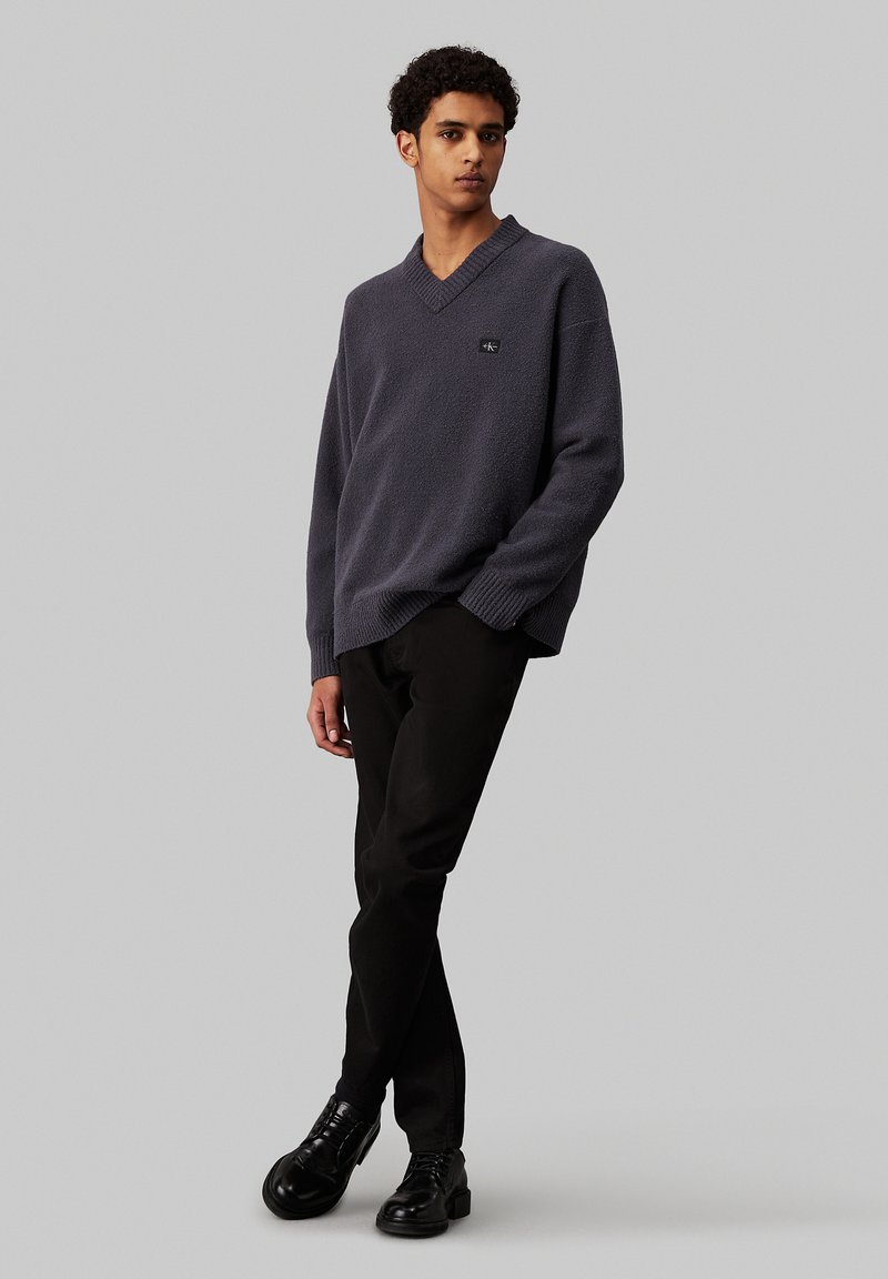 Suéter de punto gris oscuro con escote en V y una sutil textura, combinado con pantalones negros ajustados y zapatos negros con cordones. Pequeño detalle del logo en el suéter.