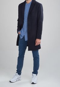 Cappotto di lana blu navy sopra una camicia azzurro chiaro con bottoni, abbinato a jeans scuri e sneakers bianche. Tessuto liscio e vestibilità su misura.