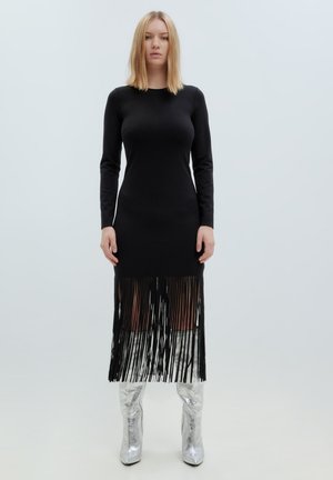 Schwarzes langärmliges Kleid mit Fransen-Saum, aus glattem Stoff. Kombiniert mit glänzenden silbernen kniehohen Stiefeln. Minimalistisches Design, ohne Muster.