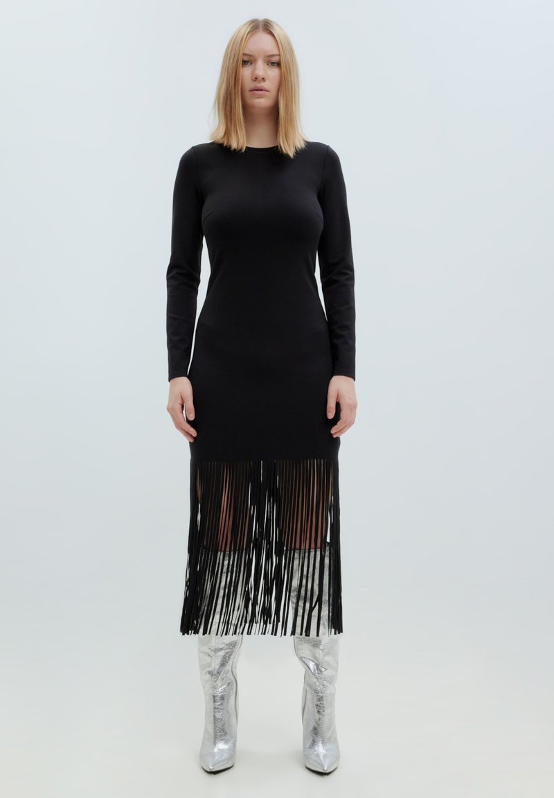 Schwarzes langärmliges Kleid mit Fransen-Saum, aus glattem Stoff. Kombiniert mit glänzenden silbernen kniehohen Stiefeln. Minimalistisches Design, ohne Muster.