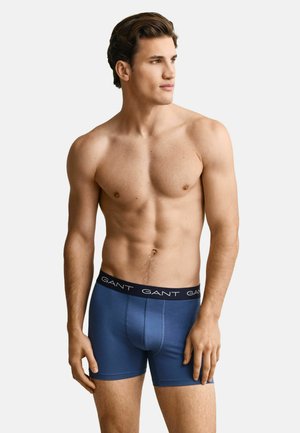 Oberkörperfreier Mann in blauen GANT-Boxershorts, steht entspannt und schaut seitlich vor einem einfarbigen Hintergrund.